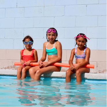 Three young girls sitting on the edge of a hot spring pool, holding an orange pool noodle toy.